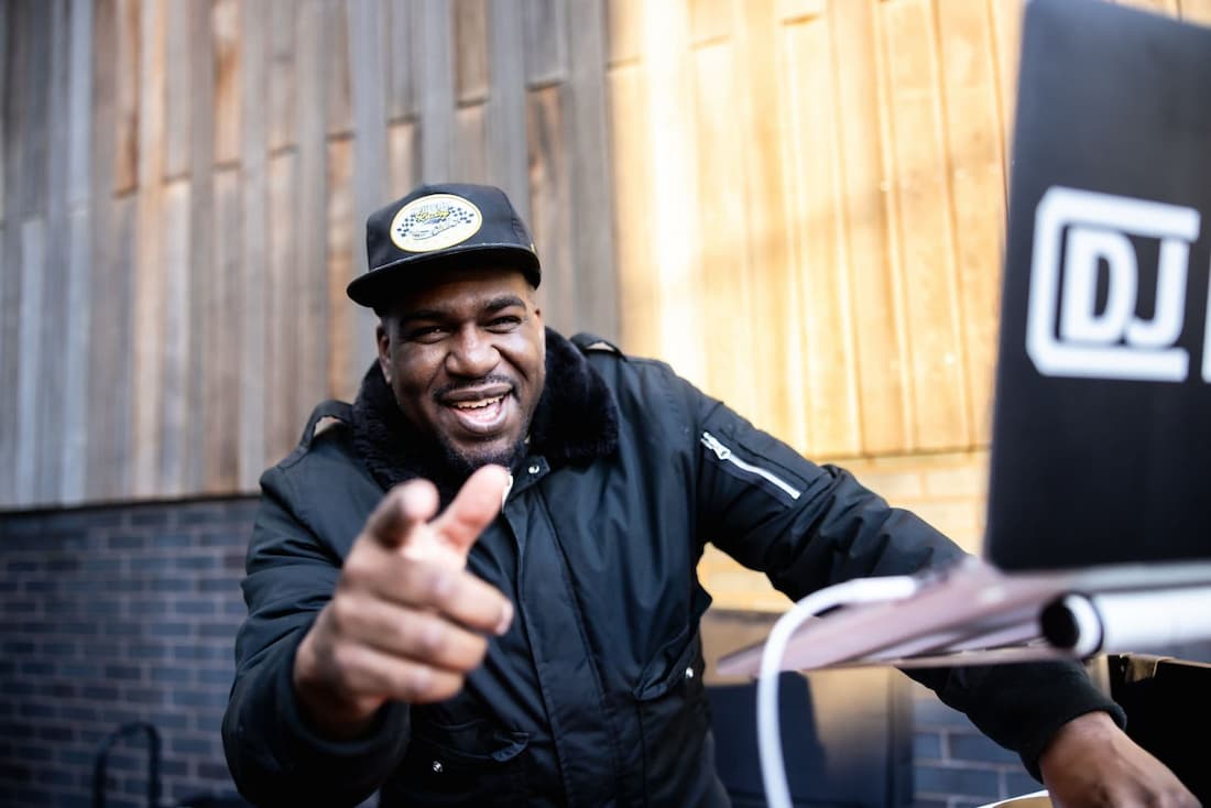 DJ Jerome wearing a black hat and jacket is pointing at the camera.