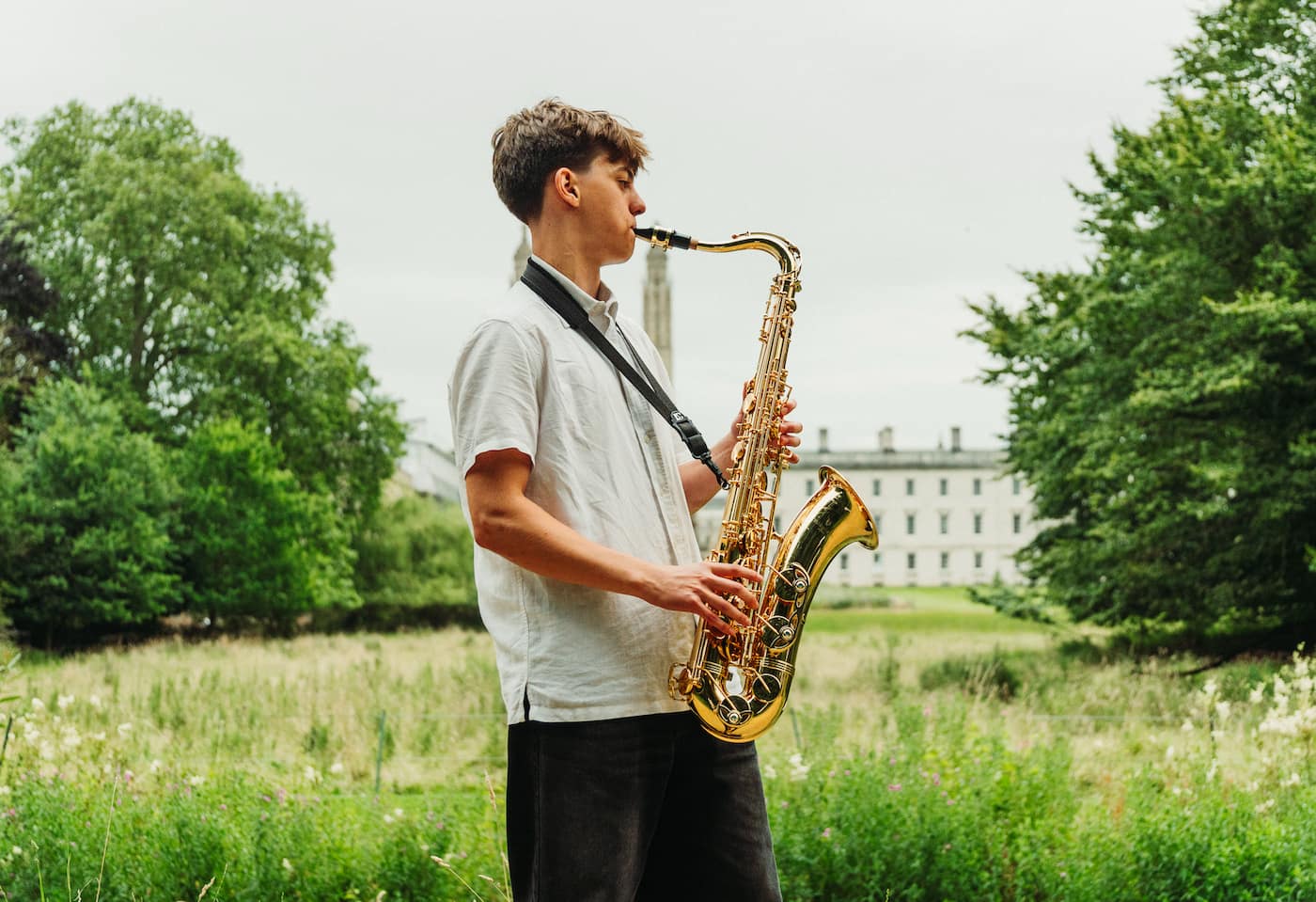 Young man playing saxophone outdoors in a lush green park with trees and a historic building in the background.