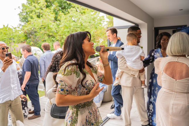 A woman holding a microphone and singing or speaking at a lively outdoor social gathering with happy guests and greenery in the background