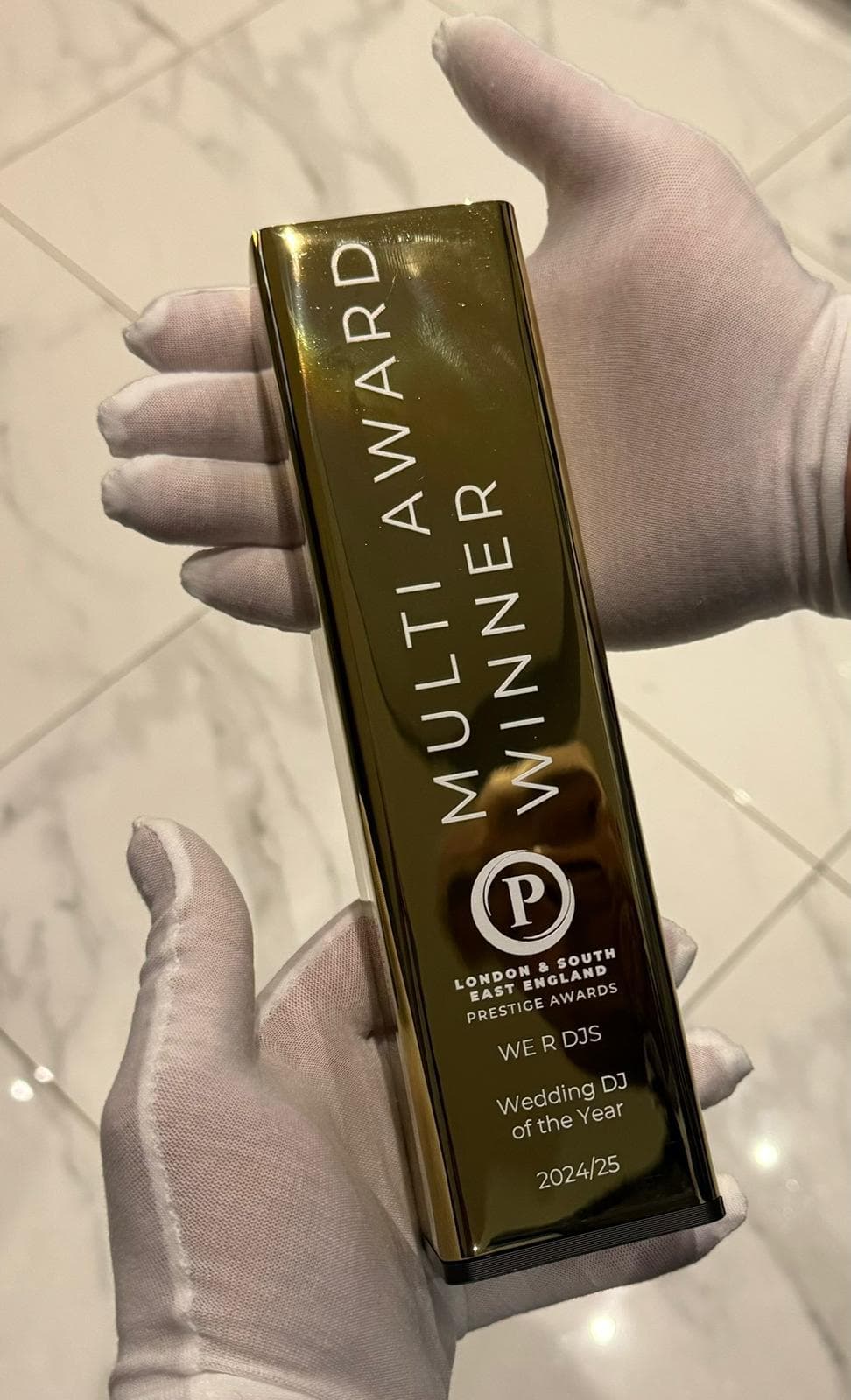 A gold award plaque that reads "MULTI AWARD WINNER," stating "WE R DJS Wedding DJ of the Year 2024/25" with "LONDON & SOUTH EAST ENGLAND PRESTIGE AWARDS" inscribed. The plaque is held in gloved hands.