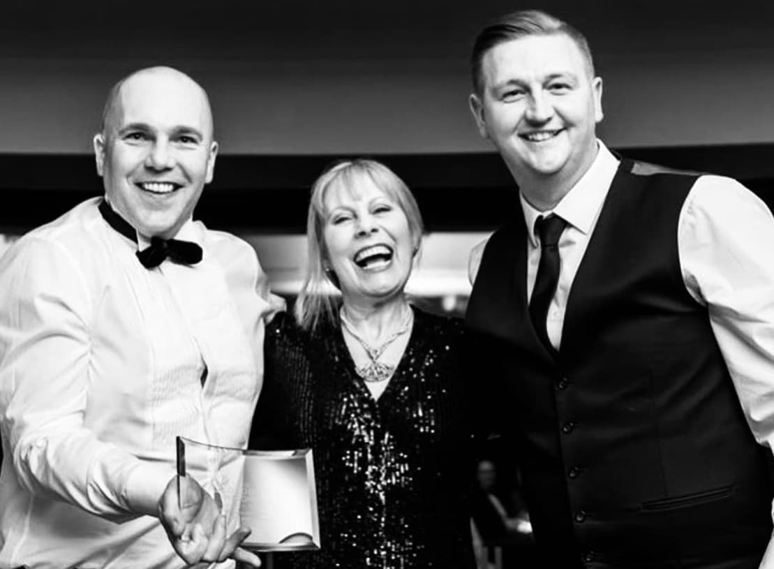 Black and white photo of three professionally dressed people smiling at a dj award event, with two men in formal attire and a woman in a sparkling dress.